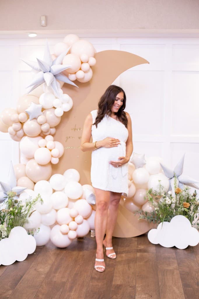 An expectant mother in white beside a crescent moon backdrop trimmed with cream balloons, silver stars and paper clouds